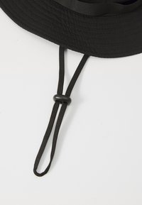 Cappello a secchiello nero con una larga visiera piatta, realizzato in tessuto testurizzato. Presenta una cintura regolabile per il mento e una corona arrotondata.