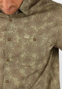 Chemise boutonnée vert olive avec un motif floral, dotée d'une poche poitrine et de boutons en bois. Tissu doux et texturé. Étiquette de marque visible.