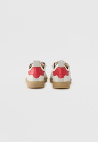 Fehér bőrből készült sneaker, bézs gumitalppal és piros bőr díszítéssel a sarkán. Cipzáras záródása és hurkos részlete van.