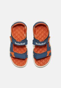 Blaue und orangefarbene Kindersandalen mit verstellbaren Riemen, texturiertem Fußbett und weißer Gummisohle. Timberland-Logo auf den Riemen.