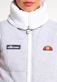 Veste grise zippée avec un col blanc haut, arborant des logos brodés sur la poitrine et un tire-zippé avec un design de logo.