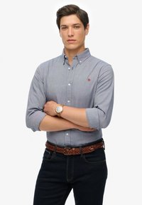 Lichtblauw knoopshirt met opgerolde mouwen, rood logo op de borst, gedragen met een donkere spijkerbroek en een bruine gevlochten riem. Draagt een horloge.