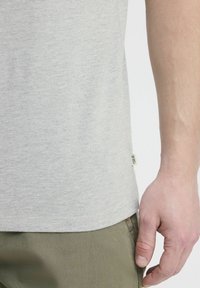 Světle šedé bavlněné tričko se spoceným střihem, krátkými rukávy a malou značkovou visačkou na boku. V páru s olivově zelenými kalhotami.