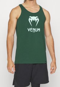 Canotta verde in cotone. Presenta una grafica di un serpente bianco e il testo "VENUM". Orlo dritto e senza maniche. Indossata con pantaloni neri.
