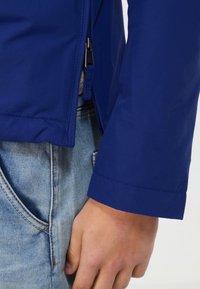 Blå nylonjacket med lynlås og rene linjer. Nærbillede af ærmet viser en glat tekstur og en lige kant. Kombineret med lys denim.