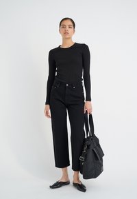Svart långärmad ribbad topp, höga svarta jeans och platta svarta skor. Bär en stor svart tote-väska med snörning upptill.