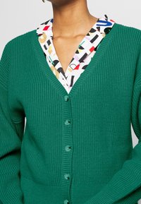 Grön ribbad kofta med V-hals, försedd med fem knappar. Under finns en mönstrad krage med olika färgglada former och mönster.