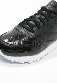 Baskets en cuir noir Reebok avec un motif géométrique en relief, lacets noirs et une semelle blanche à crampons, visibles sur un fond blanc.