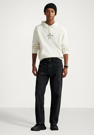 Felpa bianca con cappuccio e tasca frontale, logo nero con scritta, abbinata a jeans neri e scarpe nere. Completa il look un berretto. Design semplice e casual.