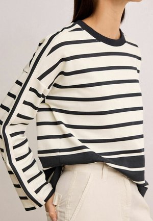 Kobieta ubrana w czarno-biały, poziomo paskowany top z długim rękawem, lekko włożony do jasnobeżowych spodni, widoczna od ramion do bioder.