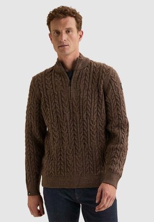 Hombre con un suéter marrón de punto trenzado con cremallera y mangas largas, y pantalones azul oscuro, de pie contra un fondo liso.