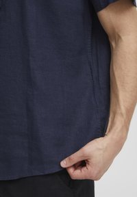 Marineblaues kurzärmeliges Shirt aus strukturiertem Stoff; mit abgerundetem Saum und einem kleinen Logo nahe des Saums. Hand greift nach dem Saum.