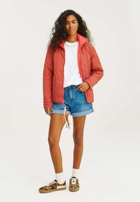 Kurtka pikowana w rdzawym kolorze z wysokim kołnierzem; połączona z białą koszulką i dżinsowymi szortami; cechuje się wzorzystymi sneakerami i podwiniętymi rękawami.