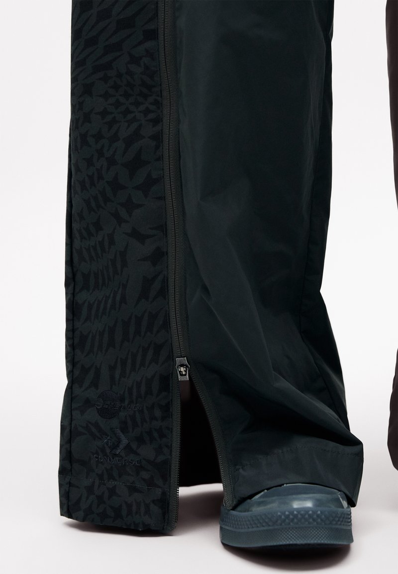 Svart vattentäta pants med ett strukturerat mönster, dragkedjestängning och en synlig blå gummisko längst ner. Har en logodetalj.