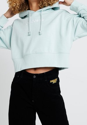 Persoon draagt een lichtblauwe, korte Adidas-hoodie en een zwart, hooggetailleerd broek met gele geborduurde details op de zak.