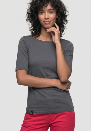 Frau mit lockigem dunklem Haar, die ein graues kurzärmliges Shirt und rote Hosen trägt, posiert mit einer Hand nahe am Gesicht und der anderen über der Taille verschränkt.