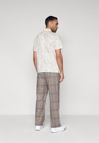 Πουκάμισο με κοντά μανίκια σε ανοιχτό μπεζ χρώμα με διακριτικά κυκλικά σχέδια, συνδυασμένο με καφέ καρό παντελόνι και λευκά αθλητικά παπούτσια. Εμφανίζεται από πίσω.