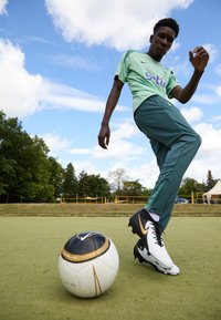 Nike futbola apavi melnbaltā krāsā ar zelta akcentiem. Spēlētājs valkā gaiši zaļu kreklu un tumši zaļas bikses. Tuvumā atrodas rakstaini futbola.