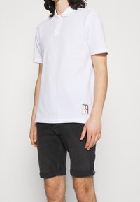 Polo shirt blanc avec un col, deux boutons et une petite étiquette avec logo en bas. Associé à un short en denim foncé retroussé.
