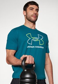 Teal bomulds t-shirt med et stort hvidt og gult Under Armour logo, kombineret med en sort kettlebell i et fitnessmiljø.