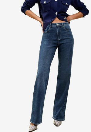 Donker gewassen denim jeans met een rechte pijp, hoge taille en subtiele stiksels. Gecombineerd met een marineblauwe button-up cardigan en gemusterde schoenen.