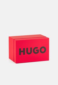 Punane ristkülikuline kast, millel on sile tekstuur, esipaneelil julge must tekst "HUGO". Minimalistlik disain, nähtavaid mustreid pole.