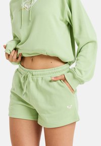 Sudadera verde claro con un logo, combinada con pantalones cortos verdes claros a juego. Ambos tienen un corte casual y un material suave y texturizado.