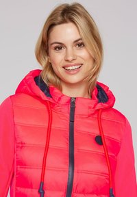 Bright pink puffer jacket with a hood, featuring navy accents, a front zipper, and drawstrings. Quilted texture and logo patch on the chest.