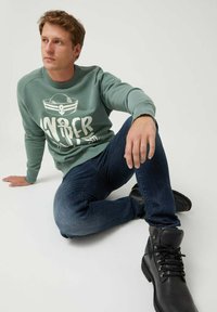 Hombre sentado en el suelo vistiendo un sudadera temática de marinero verde, jeans oscuros y botas de arriba negras, mirando hacia la izquierda.