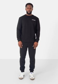 Set di felpa nera e pantaloni sportivi. La felpa presenta un collo rotondo e un logo sul petto; i pantaloni hanno polsini elastici. Completato con scarpe da ginnastica bianche.