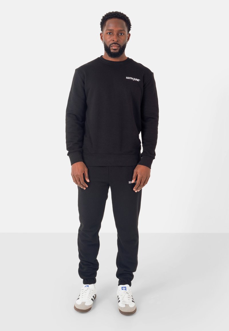 Set di felpa nera e pantaloni sportivi. La felpa presenta un collo rotondo e un logo sul petto; i pantaloni hanno polsini elastici. Completato con scarpe da ginnastica bianche.