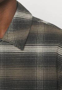 Rutig skjorta med en färgpalett i mörk och ljusgrå, med en strukturerad krage och mjuk tygtextur. Designen inkluderar varierande rutmönster.