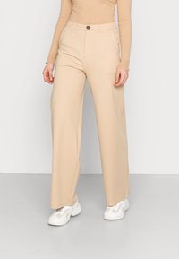 Femme portant un pantalon large beige, un haut à manches longues beige côtelé et des baskets blanches à semelle épaisse, se tenant sur un sol gris.