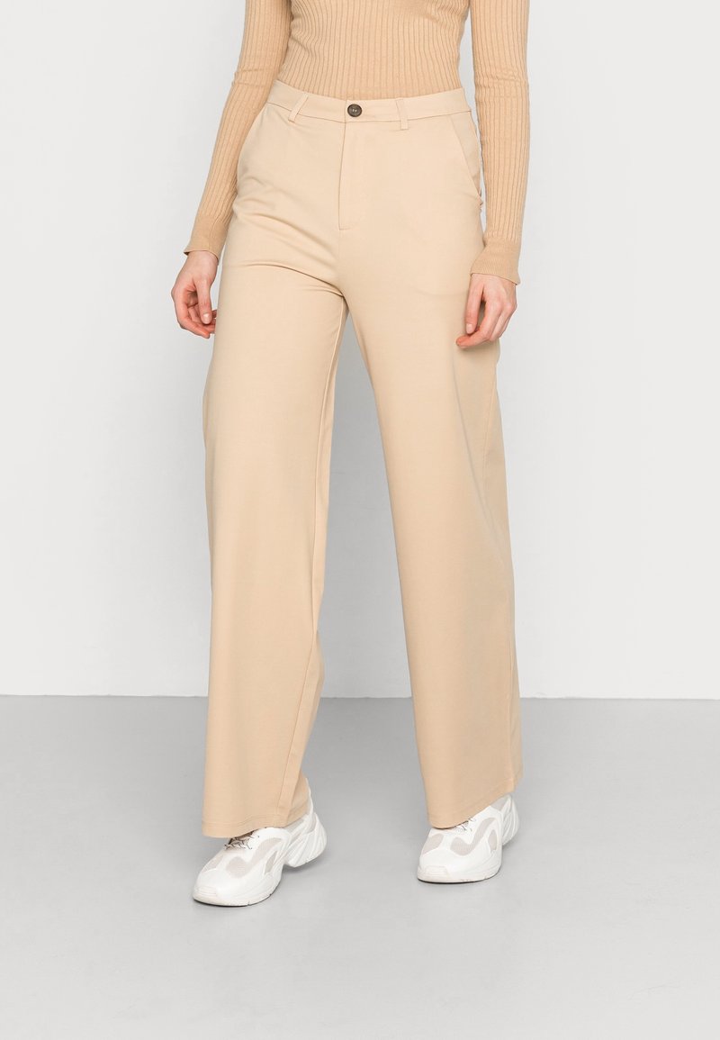 Femme portant un pantalon large beige, un haut à manches longues beige côtelé et des baskets blanches à semelle épaisse, se tenant sur un sol gris.