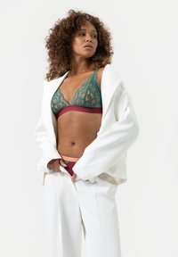 Groene kanten bralette met een bordeauxrode band, gecombineerd met bijpassende bordeauxrode slips. Model draagt een oversized witte blazer. Gladde texturen.