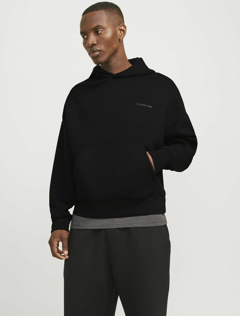 Sweatshirt à capuche noir avec une poche avant et un ourlet côtelé. Présente un logo discret sur la poitrine et est associé à un pantalon sombre. Design simple et décontracté.