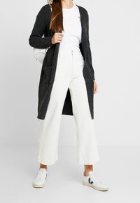 Ciemnoszary długi kardigan z otwartym przodem, z dwoma kieszeniami, noszony na białej koszulce i białych szerokich spodniach z sztruksu. Stylizowany z białymi trampkami.