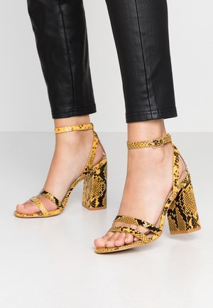 Żółto-czarne sandały na obcasie w wzór węża, z blokowym obcasem, regulowanym paskiem na kostkę i wieloma cienkimi paskami na stopie.