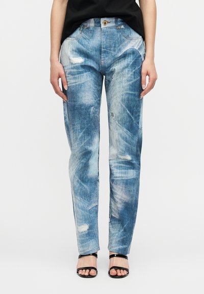 Femme portant un jean droit bleu clair déchiré et des sandales noires à bout ouvert, debout avec les bras relâchés le long du corps.