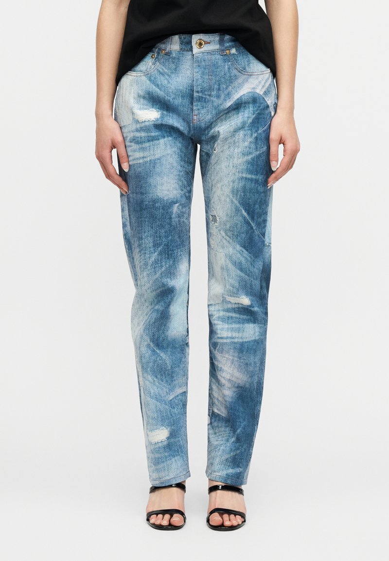 Γυναίκα με ανοιχτό μπλε τζιν παντε λόνι με φθορές και μαύρα σανδάλια με ανοιχτά δάχτυλα, στέκεται με τα χέρια χαλαρά στο πλάι.