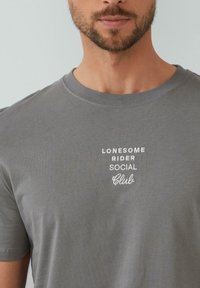 Szara bawełniana koszulka z krótkim rękawem, z białym haftowanym napisem: "LONESOME RIDER SOCIAL Club" w okolicy kołnierza. Miękka faktura.