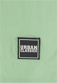 Világoszöld anyag, texturált felülettel, fekete négyzet alakú címkével, amelyen fehér, vastag betűkkel szerepel az "URBAN CLASSICS" felirat.