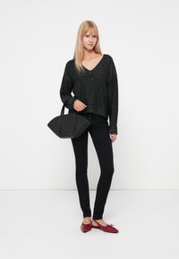 Maglione nero a maglia con scollo a V, abbinato a jeans skinny neri e scarpe basse rosse. La modella tiene in mano una borsa nera strutturata.