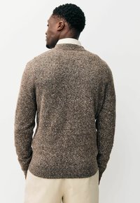 Brauner Strickpullover mit strukturiertem Muster, rundem Ausschnitt sowie gerippten Bündchen und Saum. Wird über einem hellen Hemd mit Kragen getragen.