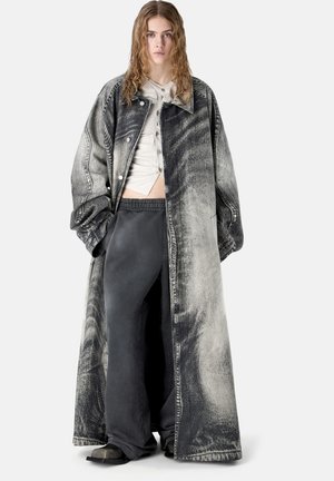 Stojící mladá žena s mokrými vlasy, oblečená v dlouhém, černo-šedě praném oversized kabátu, zapnutém béžovém topu, volných černých kalhotách a černých botách.