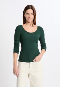 Kobieta z prostymi brązowymi włosami, ubrana w dopasowaną ciemnozieloną bluzkę z długim rękawem i jasnobeżowe spodnie, stojąca z jedną ręką w kieszeni.