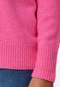 Pull en tricot rose avec un poignet et un ourlet côtelés, arborant une texture légèrement pailletée. Gros plan sur la manche révélant le motif tricoté.