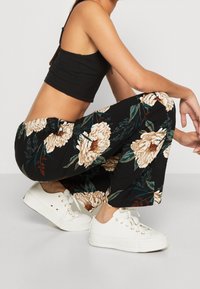 Kobieta w przysiadzie, ubrana w czarny top crop, czarne szerokie spodnie z dużym białym nadrukiem w kwiaty oraz białe sneakersy na sznurówki.