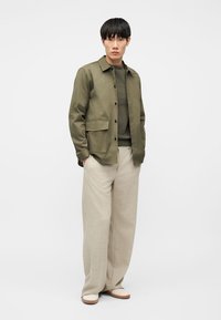 Mann trägt olivgrüne, geknöpfte Jacke über dunkelgrünem Pullover, beige Hosen mit weitem Bein und weiße Schuhe, steht mit Händen in den Taschen.