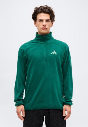 Jeune homme debout, portant une polaire verte à fermeture éclair quart avec un petit logo triangulaire sur la poitrine gauche, et un pantalon noir.
